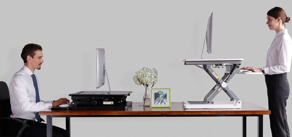 Zit-sta werkplek in actie: links zittend en rechts staand met de Adjustable Sit-Stand Desk Riser 2. Twee mensen werken met de Adjustable Sit-Stand Desk Riser 2: links zittend en rechts staand, wat het zit-sta principe illustreert.
